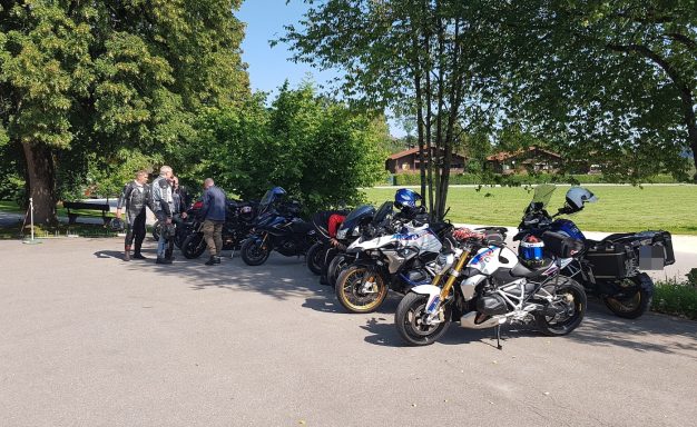 Neun Motorräder parken auf einem Parkplatz, Fahrer stehen daneben in einer grünen schattigen Umgebung.