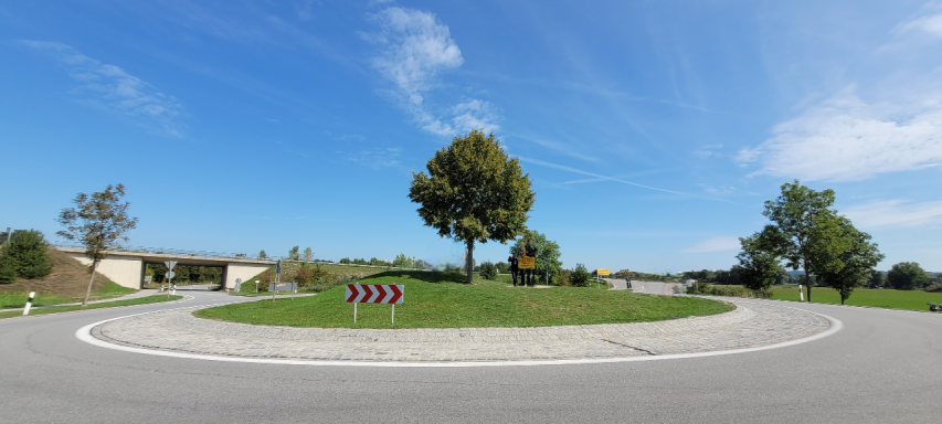 Kreisel mit Grünfläche und Bäumen unter blauem Himmel und wenigen Wolken.