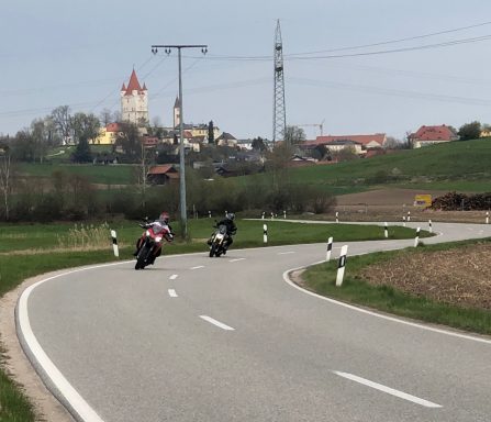 Zwei Motorräder auf einer kurvigen Landstraße mit Blick auf ein Schloss im Hintergrund.