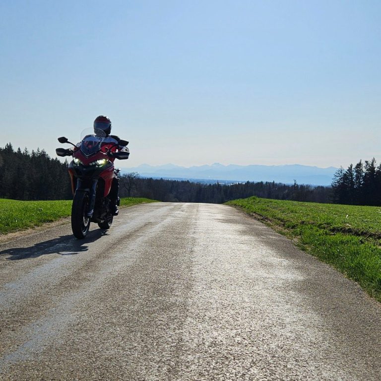 Ein Motorrad steht auf einer schmalen, sonnigen Straße mit Grünflächen und die Chiemgauer Alpen im Hintergrund.