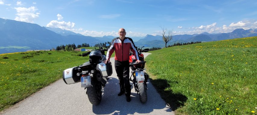 Motorradfahrer steht auf der Straße mit zwei Motorrädern vor einer Alpenlandschaft.