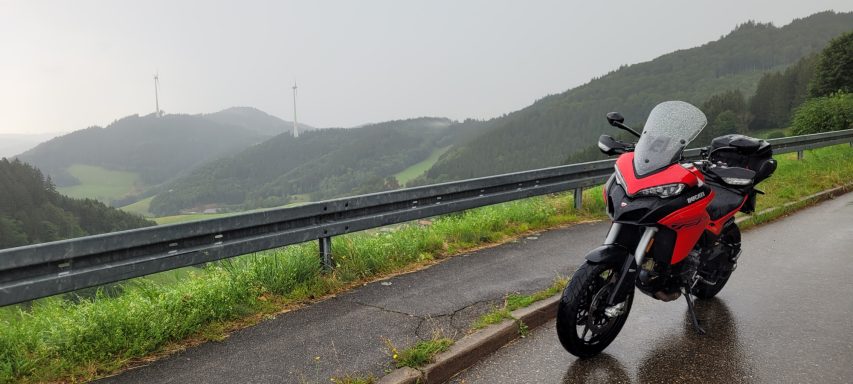 Rotes Motorrad auf einer nassen Straße mit grünen Hügeln im Hintergrund.
