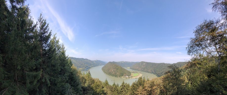 Panoramablick auf einen Fluss, umgeben von bewaldeten Hügeln und blauem Himmel.