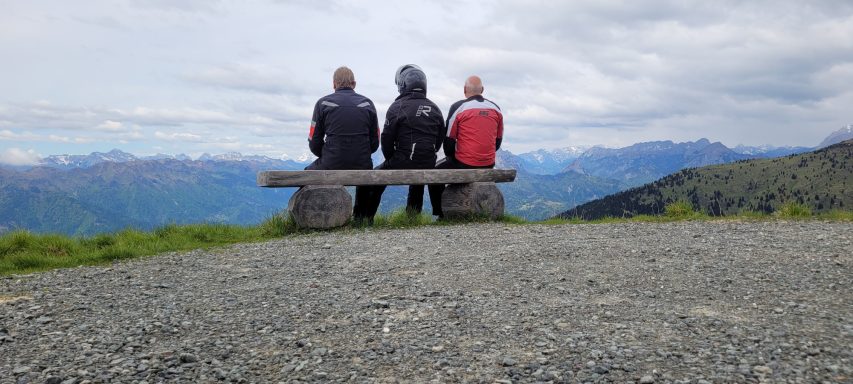 Drei Personen sitzen auf einer Bank mit Aussicht auf Berglandschaft.