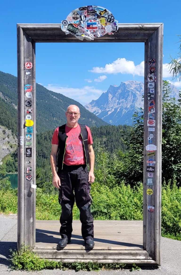 Mann mit rotem Shirt und schwarzer Weste steht in einem Rahmen vor der Zugspitze.