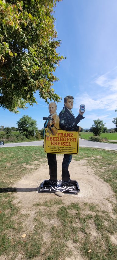 Zwei Personen stehen unter einem Baum, mit einer Werbetafel auf der Wiese.