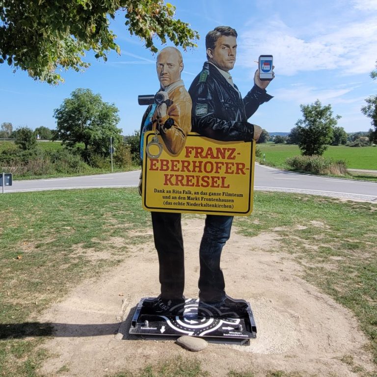 Eberhofer Kreisel Zwei lebensgroße Figuren stehen für den "Franz-Eberhofer-Kreisverkehr" in einer Landschaft.