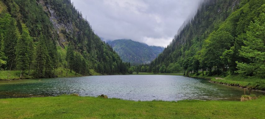 Panorama eines ruhigen Sees umgeben von grünen Bergen und bewaldeten Hängen.