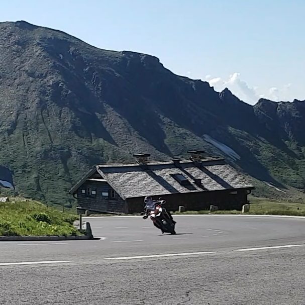 Ein Motorrad fährt durch eine Kehre auf einer sonnigen Straße mit Grünflächen und den Alpenhauptkamm im Hintergrund.